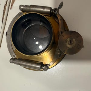 Könnte beinhalten: Ein antiker Kameraverschluss-Mechanismus aus Messing mit einer zentralen kreisförmigen Linsenöffnung, metallisch silbernen zylindrischen Komponenten und einem runden Einstellrad mit eingravierten Zahlen wie 25 und 100 sowie einer gebogenen Skala.