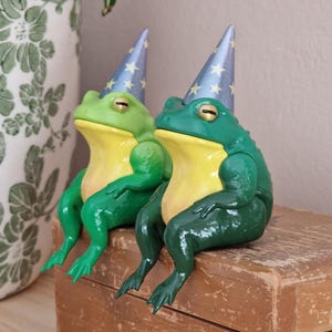 May include: Two sitting frog figurines, one in a lighter green and one in a darker green, both featuring yellow bellies and wearing pointed blue party hats decorated with small yellow stars. The frogs have smooth, glossy skin and gold-colored eyes.