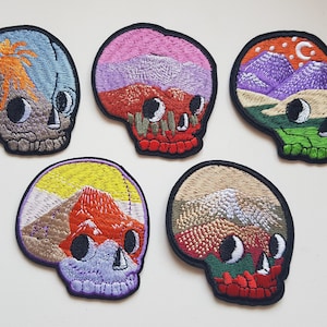Peut inclure: Cinq patchs brodés en forme de crâne avec différents motifs de paysages. Les crânes sont noirs avec des yeux blancs et sont remplis de paysages colorés. Les paysages comprennent des montagnes, un volcan et un désert.