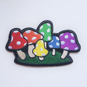May include: An embroidered patch featuring five colourful mushrooms with white polka dots. The mushrooms are arranged in a cluster on a green background.