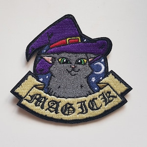 May include: Iron-on patch featuring an embroidered grey cat wearing a purple witch's hat with a red band. The cat is surrounded by a yellow banner with the word "MAGIC" in black lettering.