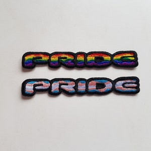 Könnte beinhalten: Zwei gestickte Aufnäher mit dem Wort "PRIDE" in Regenbogen- und Transgender-Flaggenfarben.