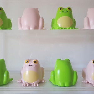 Op de afbeelding: Acht kikkerfiguurtjes, vier groen en vier roze, gerangschikt in twee rijen op een wit rek. De kikkers kijken in verschillende richtingen, sommige kijken naar voren, sommige kijken naar de zijkant en sommige kijken naar achteren.