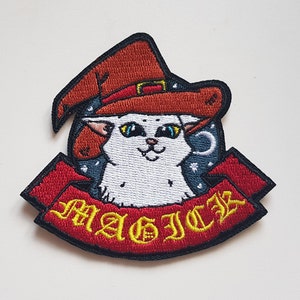 May include: White cat wearing a red and brown witch hat with a black brim. The cat is surrounded by a blue background with stars and a crescent moon. A red banner with the word "MAGIC" in yellow letters is below the cat.