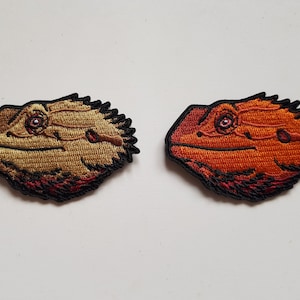 May include: Two embroidered patches of a bearded dragon's head. One is golden brown with red details, the other is orange with red. Both have black outlines and detailed stitching.
