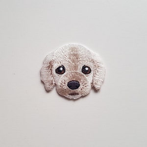 May include: Embroidered patch of a white dog with brown eyes and a black nose. The dog is looking directly at the viewer.