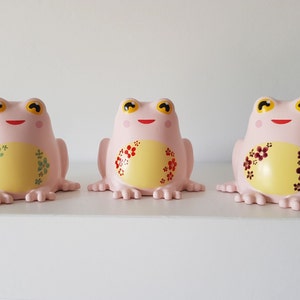 Pode incluir: Três estatuetas de sapo de cerâmica rosa com barrigas amarelas e desenhos florais pintados. Os sapos têm olhos pretos e bocas vermelhas.
