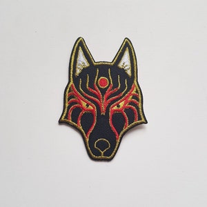 以下が含まれることがあります： 黒と金の刺繍が施された狼のマスクのパッチ。赤いアクセントと中央に白い円があります。