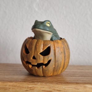 May include: A small, dark green frog with bright yellow eyes sits inside a carved orange pumpkin. The pumpkin features a classic jack-o'-lantern face with triangular eyes and a jagged mouth, showcasing a textured, weathered surface finish.