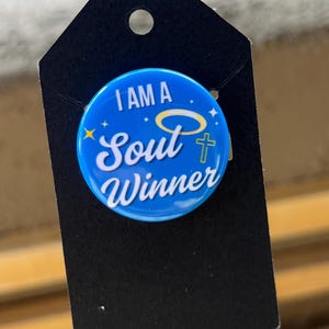 May include: A blue button with the text "I AM A Soul Winner" in white, with a halo and cross design. The button is attached to a black tag with a hole at the top.