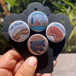 May include: A set of four round Chicago-themed buttons. One button features "The Bean" sculpture, another the Chicago skyline, a third a pizza, and the last a Chicago Bears logo. The buttons are attached to a black card.