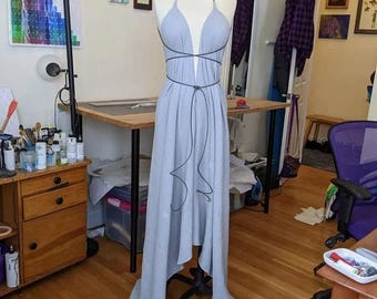 May include: A light blue floor-length dress on a mannequin, featuring a deep V-neckline, thin halter straps, and a wrap-around cord detail that ties at the waist. The fabric has a subtle texture, and the skirt has an asymmetrical hemline.