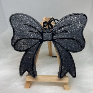 May include: A black glitter bow with a black ribbon, displayed on a wooden easel.