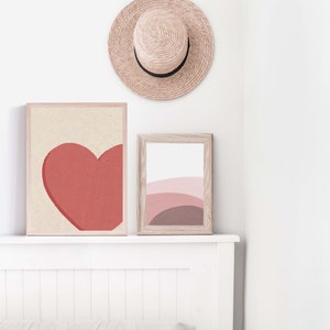 May include: Two framed abstract art prints on a white wall above a white headboard. The print on the left features a red heart shape. The print on the right features a pink and brown abstract design. A straw hat hangs on the wall above the prints.