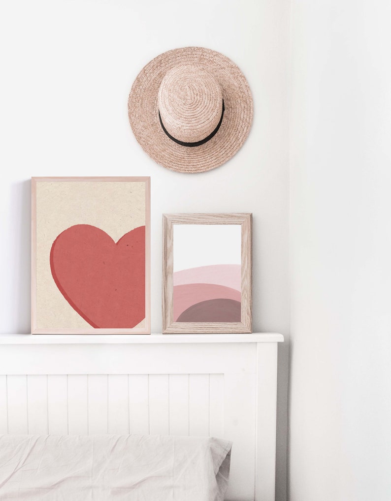 May include: Two framed abstract art prints on a white wall above a white headboard. The print on the left features a red heart shape. The print on the right features a pink and brown abstract design. A straw hat hangs on the wall above the prints.