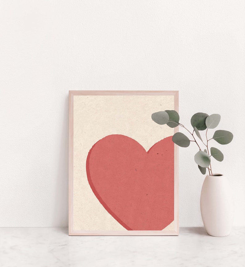 May include: A framed print of a simple, minimalist design featuring a large, coral-colored heart on a cream background.