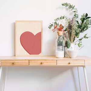 May include: A wooden desk with three drawers and white metal legs. A framed print with a red heart on a cream background sits on the desk. A glass vase with dried flowers and a white bowl are also on the desk.