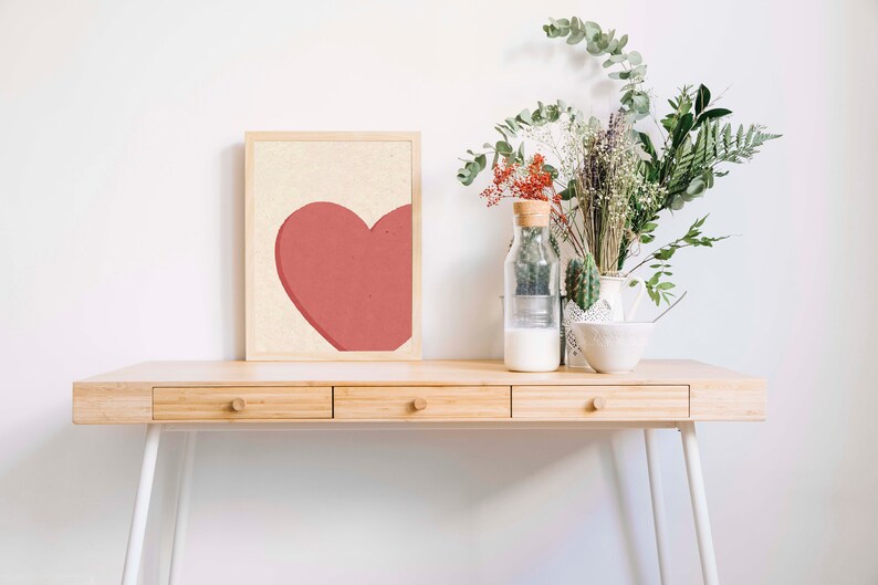 May include: A wooden desk with three drawers and white metal legs. A framed print with a red heart on a cream background sits on the desk. A glass vase with dried flowers and a white bowl are also on the desk.