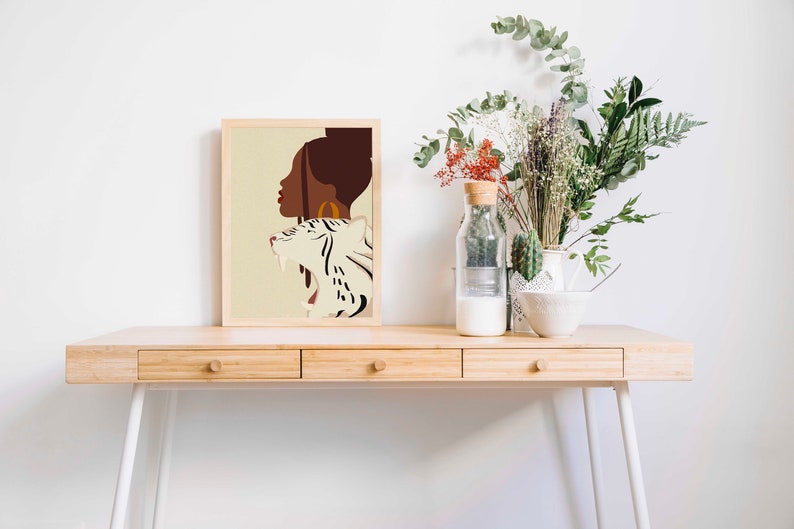 Pu&ograve; includere: Una stampa incorniciata di un'illustrazione digitale di una donna con una tigre bianca. La donna ha la pelle marrone e un orecchino a cerchio dorato. La tigre &egrave; bianca con strisce nere. Lo sfondo &egrave; di colore beige chiaro.