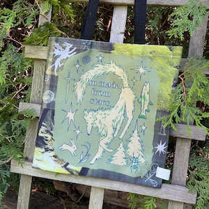 May include: A canvas tote bag with a green and black background. The bag features a light green central design with a wolf, stars, a sword, and the text "I'm made from stars". The bag has black handles.