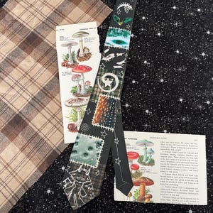 Peut inclure: Une cravate noire avec un motif patchwork représentant une étoile blanche, un œil vert, un squelette et une créature blanche ailée. La cravate est posée sur une surface à motifs noirs et blancs avec deux livres ouverts montrant des illustrations de champignons.