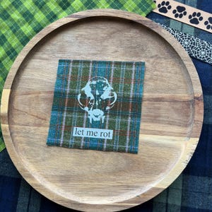 May include: A square fabric patch featuring a green, blue, and brown plaid pattern. The patch displays a white graphic of a skull and the text "let me rot" in a simple font. The patch is centered on a round, light brown wooden serving tray.