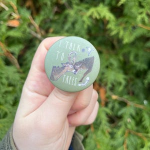 Puede incluir: Un botón verde con un ciervo blanco y el texto "I talk to trees" en blanco. También hay una estrella, un círculo y una espina de pescado en el botón.