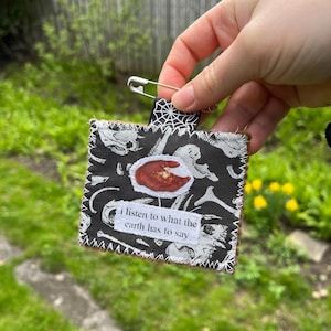 May include: A small, square fabric item with a black background featuring white bone and skull designs. A red and brown image is centered on the fabric, with the text "i listen to what the earth has to say" below. A black and white spiderweb ribbon is attached with a safety pin.