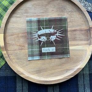 May include: A square fabric patch featuring a green, brown, and blue plaid pattern. The center displays a white graphic of a spiked collar with the text "fuck ai" printed in a simple, lowercase sans-serif font directly below the collar.