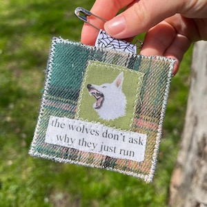 Könnte beinhalten: Ein quadratischer Stoffflicken mit einem grün-braunen Karomuster. Ein kleineres grünes Quadrat zeigt eine weiße Wolfsillustration. Der Text "the wolves don't ask why they just run" ist darunter gedruckt. Eine Sicherheitsnadel ist befestigt.