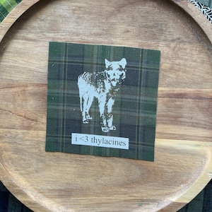 Pode incluir: Um emblema de tecido quadrado com um padrão xadrez em verde escuro e preto. Centrada no emblema está uma ilustração gráfica branca de um tilacino. Abaixo do animal, uma etiqueta retangular branca exibe o texto preto i <3 thylacines.