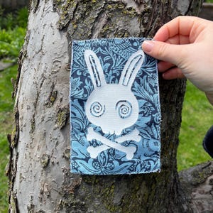 May include: A blue fabric patch with a white rabbit skull and crossbones design. The background features a floral pattern in shades of blue. The patch is held against a tree trunk.