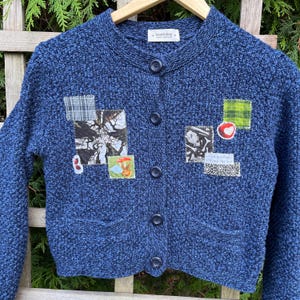 Peut inclure: Un cardigan en tricot bleu avec une variété de patchs en tissu cousus sur le devant. Les patchs comprennent un motif à carreaux, un arbre, un champignon, un lapin, un cœur et un morceau de dentelle avec le texte "Je grandis lentement au soleil".