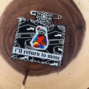 Peut inclure: Épingle à tissu faite à la main avec un fond noir représentant un ours en peluche en manteau rouge et membres colorés. L'épingle porte l'inscription "I'll return to moss" et un motif de toile d'araignée. Une épingle de sûreté argentée est attachée.