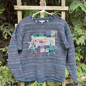 Può includere: Un maglione blu a maniche lunghe con un motivo a maglia testurizzato caratterizzato da strisce orizzontali rosse e grigie. La parte anteriore è decorata con un design patchwork di tessuto scozzese, una forma di pesce e stelle, con vari bottoni.