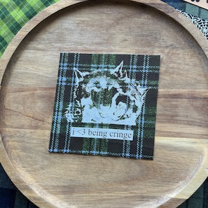 May include: A square fabric patch featuring a dark green, black, and light blue plaid pattern. The center displays a white graphic of a howling wolf with the text "i <3 being cringe" printed in a simple black font on a white rectangular background.