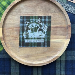 May include: A square fabric patch featuring a dark green, black, and blue plaid pattern. The patch displays a white graphic of a wolf head with its mouth open and the text "i'll eat you whole" printed in white letters on a rectangular background.