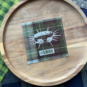 May include: A square fabric patch featuring a green, brown, and blue plaid pattern. The center displays a white graphic of a spiked collar with the text #SWAG printed in white block letters directly below the collar illustration on the fabric.