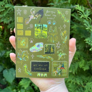 Peut inclure: Une planche d'autocollants rectangulaire vert olive avec diverses illustrations holographiques, dont un scarabée, un crâne, un squelette de poisson et des forêts. Le texte inclut "do you hear the trees?", "I love touching grass" et "We are all connected".