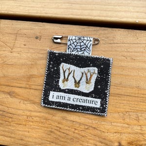May include: A fabric pin with a black background and white stars. The pin features a white rectangle with three antler illustrations and the text "i am a creature". A small fabric piece with a spiderweb design is attached to a safety pin.