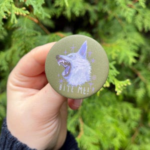 Könnte beinhalten: Ein runder, olivgrüner Knopf mit einer Illustration eines knurrenden Wolfs in Blau- und Weißtönen. Der Text "BITE ME!" steht in Weiß unter dem Wolf. Der Knopf wird vor einem Hintergrund aus grünem Laub gehalten.