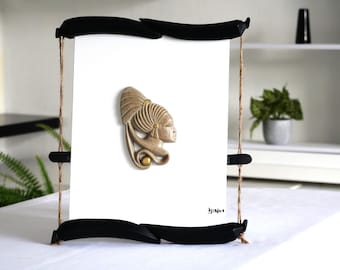 May include: A beige and gold ceramic sculpture of a woman's head with a decorative headdress. The sculpture is framed in a black wooden frame with a string hanging from the top.