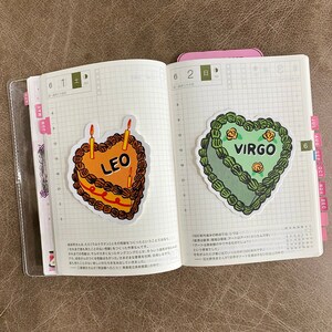 May include: Two heart-shaped birthday cakes with candles, one decorated in orange and brown with the word "LEO" and the other in green with the word "VIRGO".