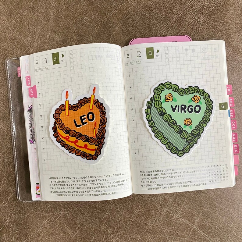 May include: Two heart-shaped birthday cakes with candles, one decorated in orange and brown with the word "LEO" and the other in green with the word "VIRGO".