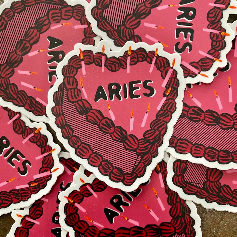 May include: A set of pink and black heart-shaped cake stickers with the word "ARIES" in black lettering. The stickers have a pink frosting design with white candles.