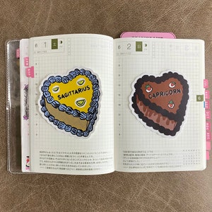 May include: Two heart-shaped cake stickers with astrological signs. The first sticker is blue and yellow with the text "SAGITTARIUS". The second sticker is brown and red with the text "CAPRICORN".