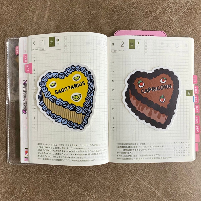May include: Two heart-shaped cake stickers with astrological signs. The first sticker is blue and yellow with the text "SAGITTARIUS". The second sticker is brown and red with the text "CAPRICORN".