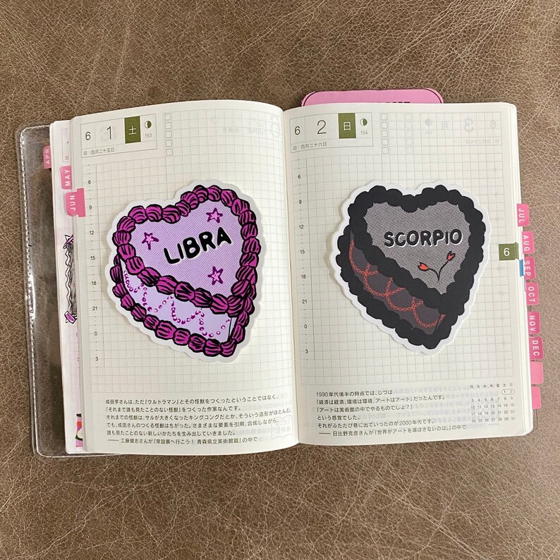 May include: Two heart-shaped stickers with astrological signs. The first sticker is pink and says "LIBRA" in white text. The second sticker is black and says "SCORPIO" in white text.