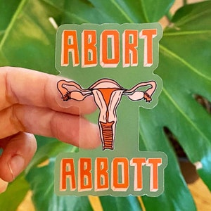 Puede incluir: Una pegatina transparente con las palabras "ABORT ABBOTT" en letras naranjas sobre un fondo verde. Una ilustración rosa y naranja de un útero se encuentra en el centro de la pegatina.