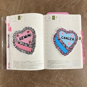 May include: Two heart-shaped cake stickers with pink and blue frosting. The pink cake has the word "GEMINI" on it and the blue cake has the word "CANCER" on it.